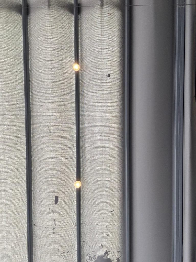 Orthographic view looking up at a grey textured fabric retractable awning canopy with integrated string light bulbs along dark metal support tracks.