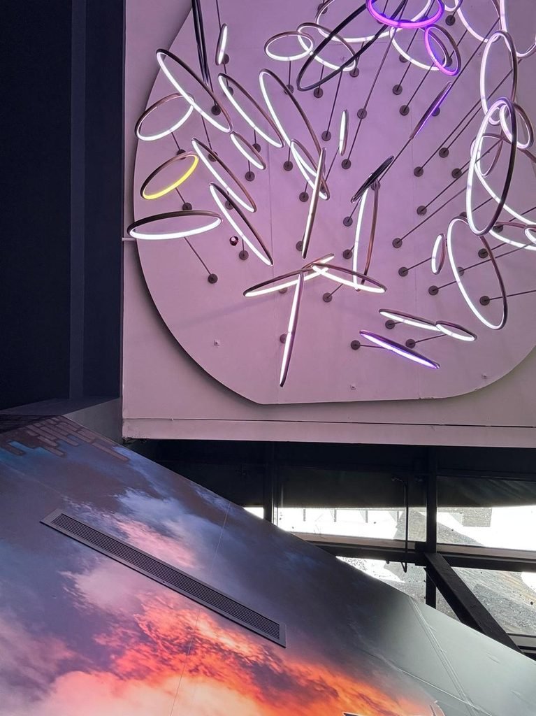 Low-angle orthographic view of a contemporary ceiling sculpture featuring angled LED ring lights suspended from a circular base plate with violet light ambient wash, adjacent to a sunset sky graphic soffit.