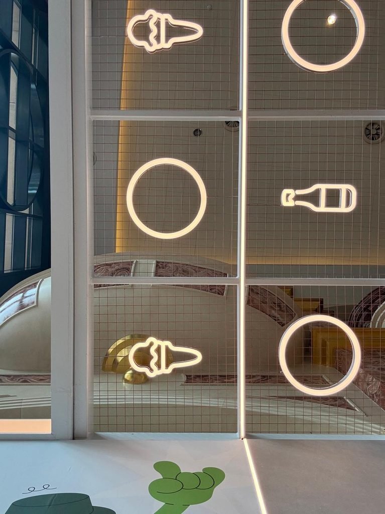 Orthographic view of a white wire mesh ceiling screen with glowing neon outline icons of ice cream cones, circles, and bottles, framed by linear LED light strips.