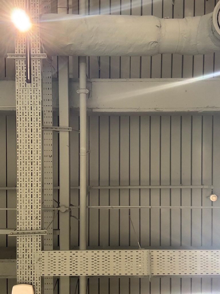 Orthographic view looking up at an industrial coffee shop ceiling with ribbed soffit decking, white painted HVAC ducts, perforated cable raceways, and a bright spotlight lens flare.