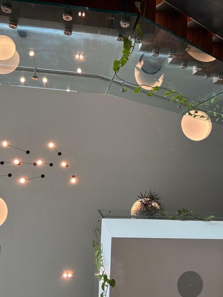 Low-angle view of a high-ceilinged hotel reception atrium featuring floating glass walkways, climbing green ivy vines, luminous globe pendant lamps, and a constellation branch chandelier fixture.