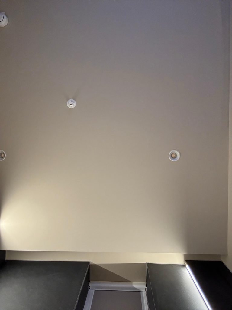 Orthographic low-angle view of a smooth taupe hotel room drop ceiling soffit with recessed downlights, smoke detectors, and a cove lighting reveal above dark headboard joinery.