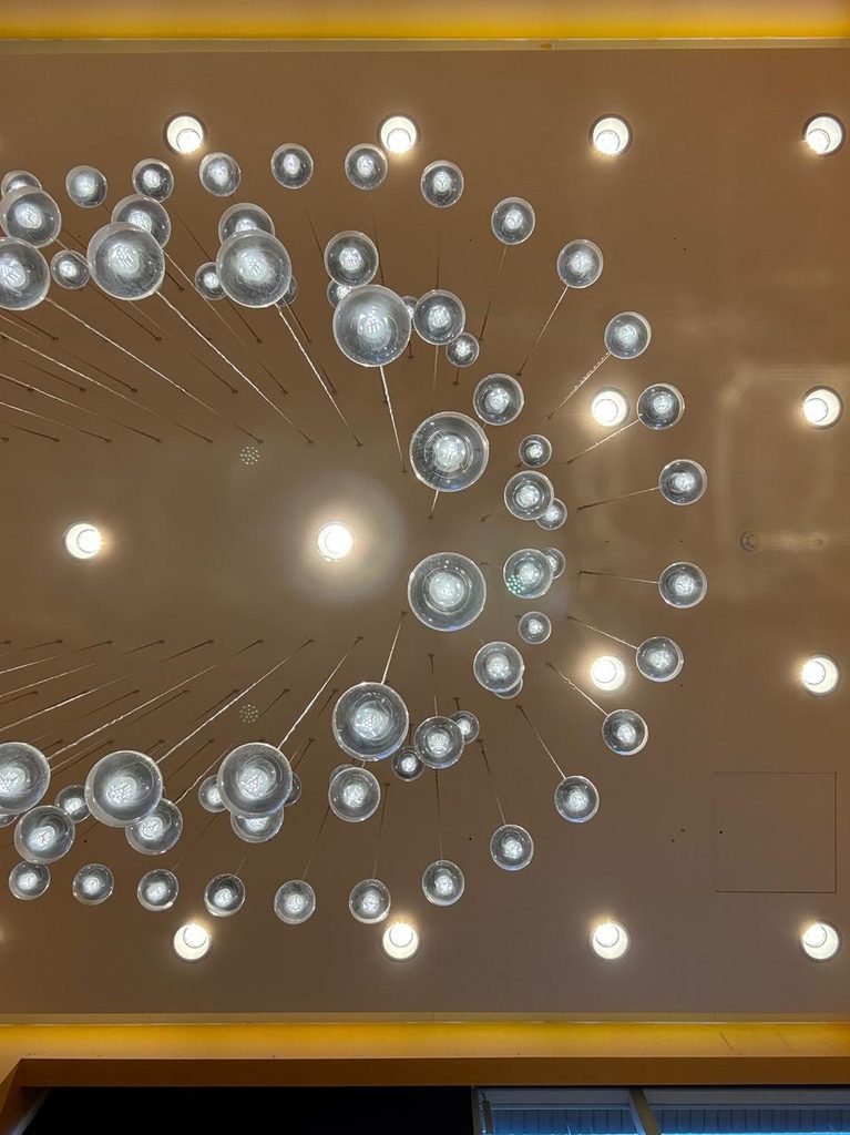 Orthographic view looking up at a warm beige plaster ceiling adorned with a radial constellation array of clear glass globe pendant lamps suspended on thin cables at cascading heights, surrounded by grid-aligned recessed downlights.