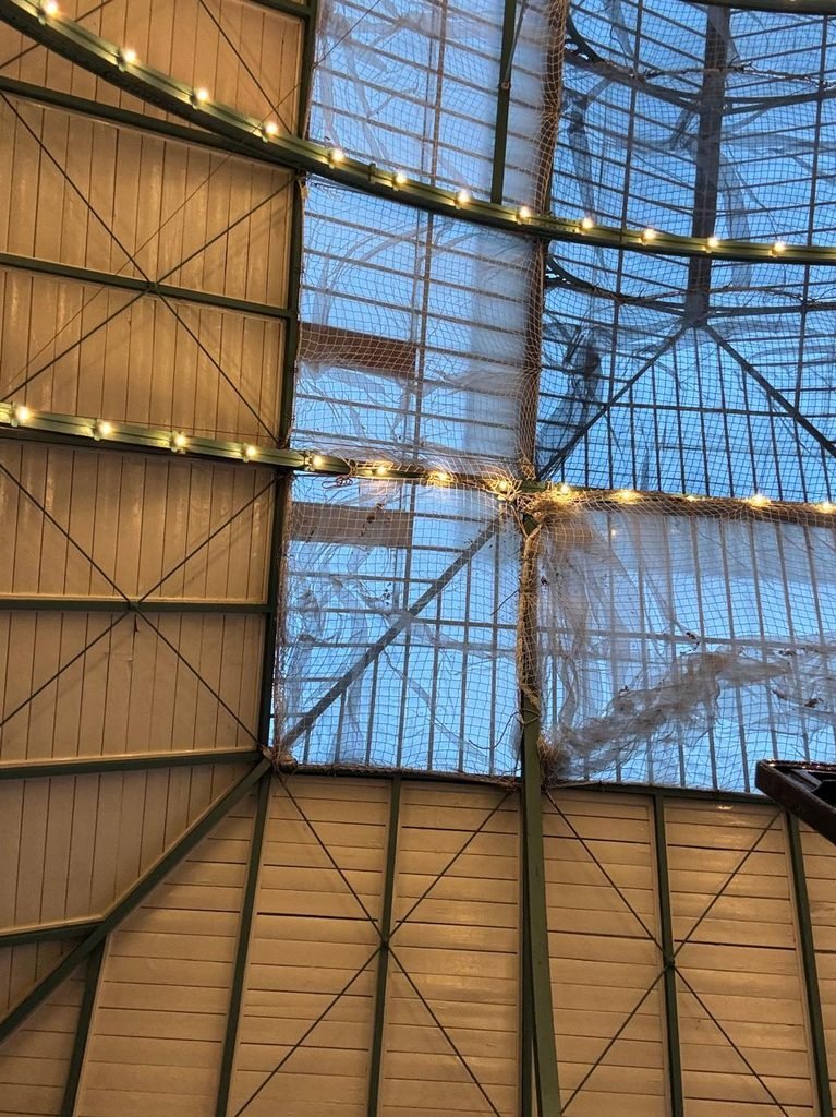 Low-angle view looking up at the historic timber and iron roof framework of Covent Garden Market, featuring green-painted iron ribs, timber tongue-and-groove decking, safety nets beneath glass panels, and string lights.