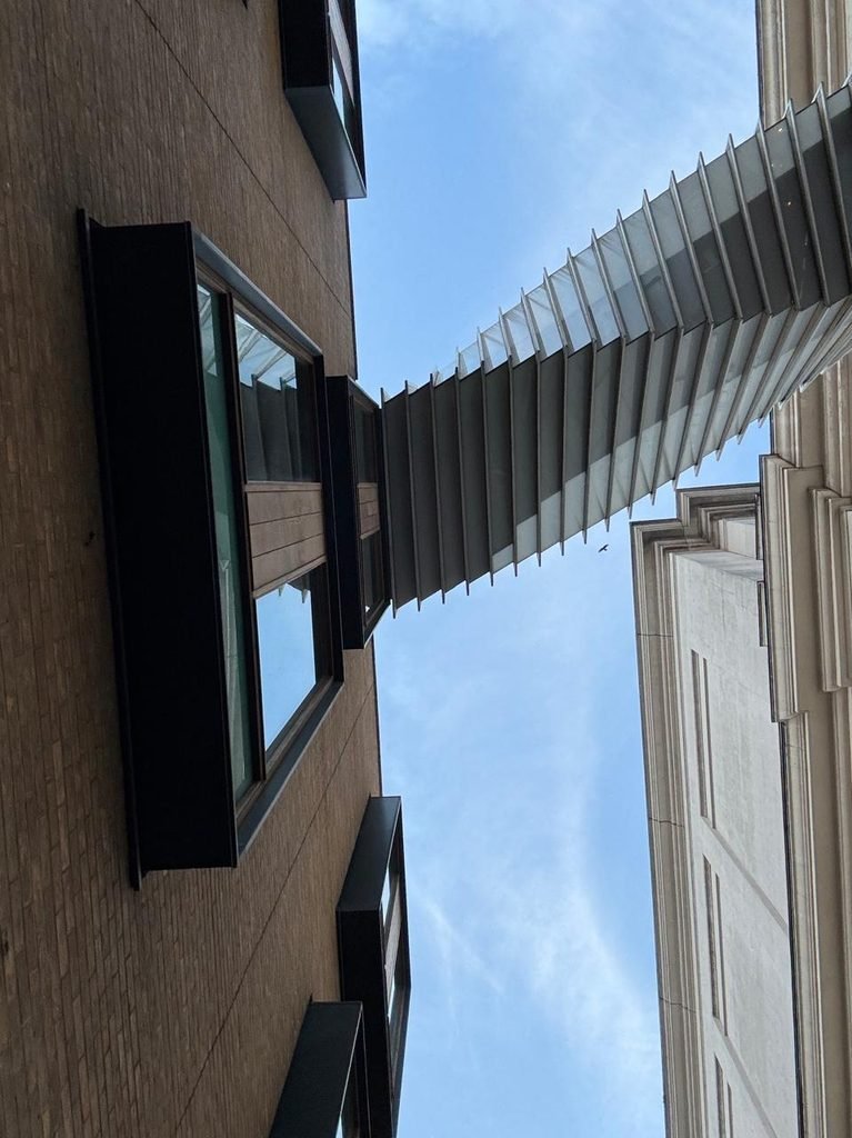 Skyward low-angle photograph looking up at the twisted concertina structural frame of the Bridge of Aspiration spanning between brick and stone building facades against a clear sky.