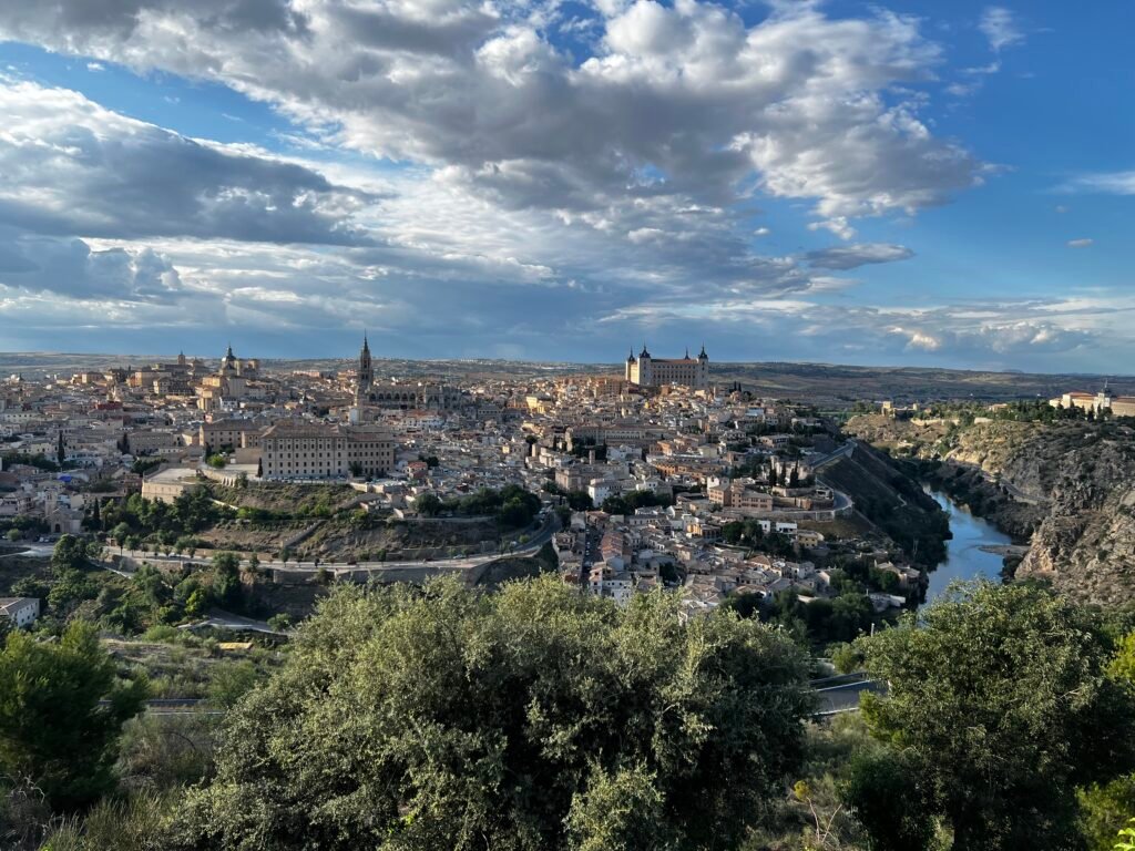 Panoramic architectural survey composite of the historic city of Toledo, Spain, capturing its medieval urban morphology, the defensive Tagus River loop, the prominent Alcázar fortification, the Gothic-Mudejar cathedral spire, and traditional stone masonry vernacular architecture integrated into rugged hillside topography.