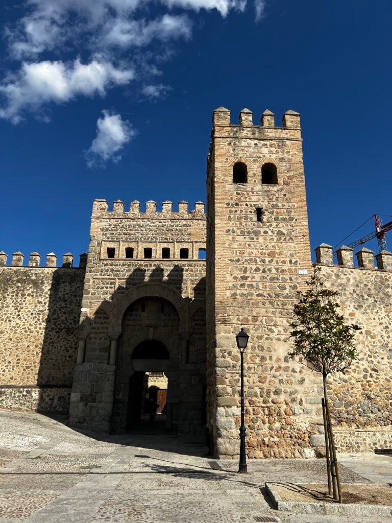 Original architectural photography of a historic fortified stone gate in Toledo, Spain, showcasing heavy structural masonry, defensive towers, and a traditional Moorish horseshoe arch characteristic of Andalusian architecture.