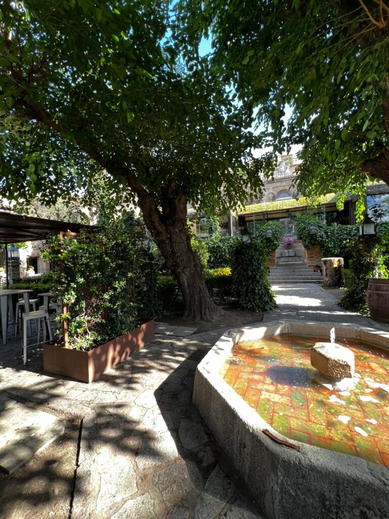 Interior view of a shaded residential courtyard in Toledo featuring a central octagonal stone fountain, mature trees, and natural stone paving, demonstrating microclimate regulation and Damascene spatial planning within Andalusian architecture.
